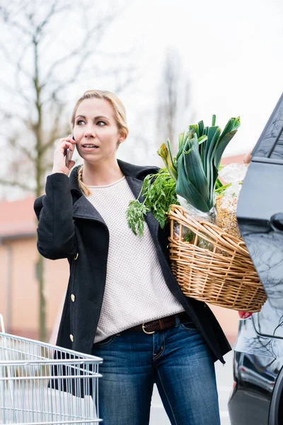 Mulher usando telefone ao armazenar mantimentos no porta-malas do carro — Fotografia de Stock