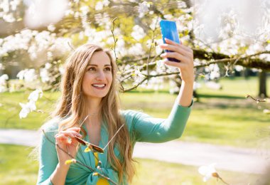 Genç bir kadın telefonuyla ağaçların çiçek açtığı parkta selfie çekiyor.