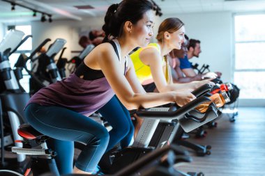 Asyalı kadın ve arkadaşları spor salonunda spor bisikletine biniyorlar.