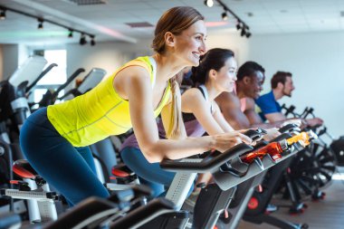 Beyaz kadın ve arkadaşları spor salonunda spor bisikletine biniyorlar.