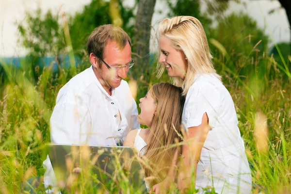 Lycklig familj sitter i äng med bärbar dator — Stockfoto