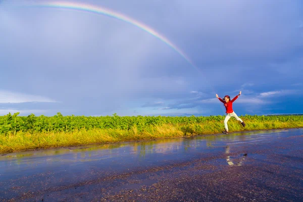 Девушка прыгает по мокрой дороге с радугой — стоковое фото
