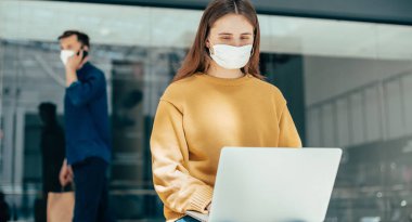 Koruyucu maskeli genç kadın sokakta dizüstü bilgisayarını kullanıyor.