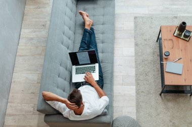 Dizüstü bilgisayarı olan genç adam kanepede yatıyor. .