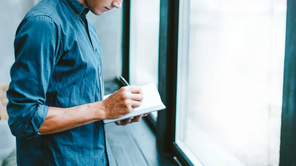 Człowiek robiący notatki w notatniku, stojący przy oknie. — Zdjęcie stockowe