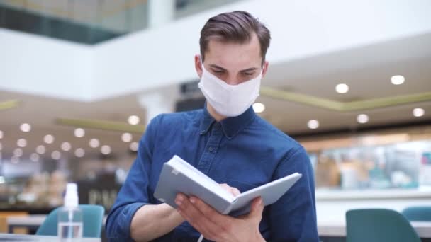 Da vicino. uomo in maschera protettiva legge un libro in un caffè . — Video Stock