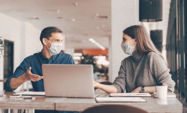 Maskeli iş arkadaşları ofis masasında oturuyorlar..