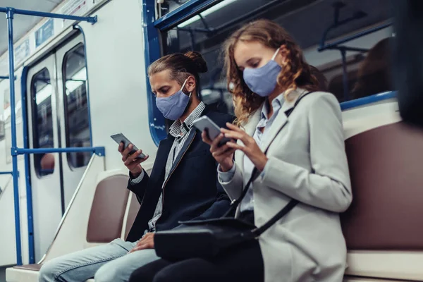 Passageiros usando máscaras protetoras usando seus smartphones enquanto sentados em um carro de metrô. — Fotografia de Stock