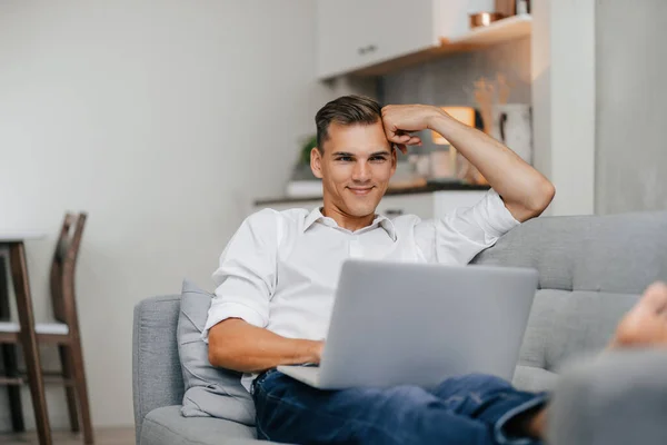 Usmívající se mladý muž procházející internet, zatímco leží na gauči. — Stock fotografie