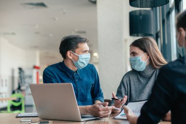 Maskeli iş arkadaşları ofis masasında oturuyorlar..