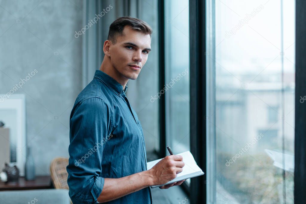 man making notes in a notebook, standing near the window.