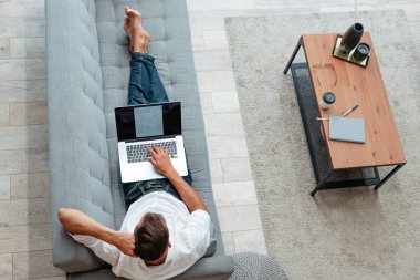 Erkek serbest çalışan, kanepede yatarken bir dizüstü bilgisayar üzerinde çalışıyor. .