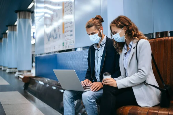 Jovem casal em máscaras protetoras usando um laptop na plataforma do metrô . — Fotografia de Stock