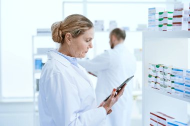İlaç dolu bir rafın yanında dijital tabletle duran kadın eczacı..