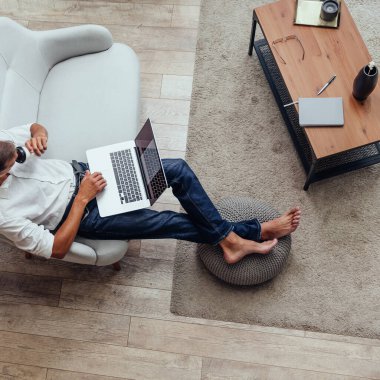 Kanepede otururken elinde bir fincan kahveyle dizüstü bilgisayar kullanan bir adam.