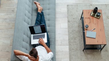 Dizüstü bilgisayarı olan genç adam kanepede yatıyor. .