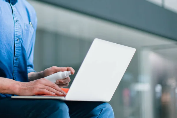 Homem com uma máscara protetora desinfecta seu laptop — Fotografia de Stock