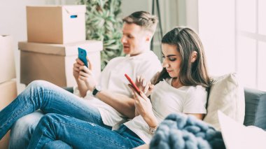 Akıllı telefonları olan genç bir çift yeni bir dairede kanepede oturuyor. .