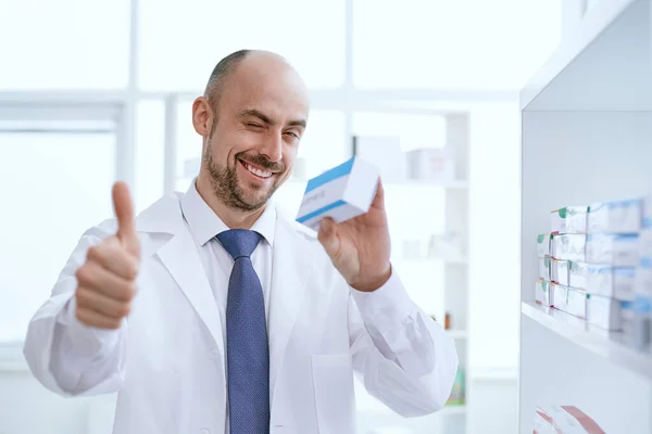 Farmacêutico sorrindo segura uma caixa de medicamento e dá um polegar para cima. — Fotografia de Stock