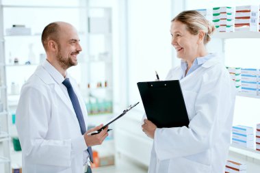 Gülümseyen eczacılar rafın yanında ilaçlarla ilgili bir şey tartışıyorlar. .