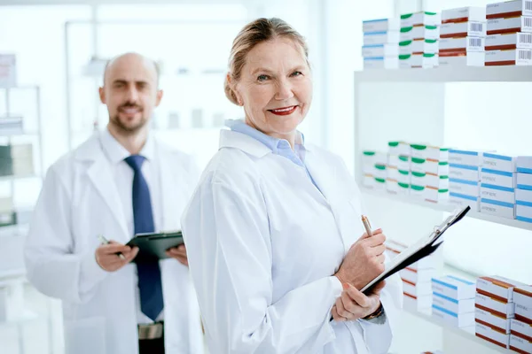 Pharmacist with a clipboard and her colleague standing near a pharmacy display case . — Stock Photo, Image