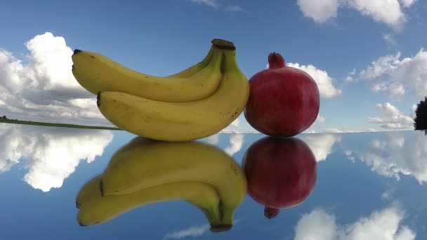 Tre bananer och ett granatäpple placeras på spegelglas under molnig himmel, tidsinställd 4k — Stockvideo
