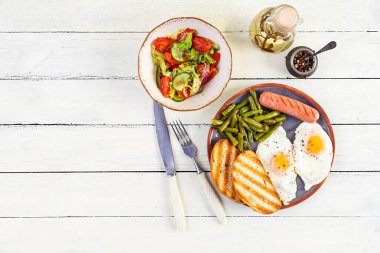 Yumurtalı kahvaltı, ızgara sosis, yeşil fasulye ve ahşap arka planda kızarmış ekmek.