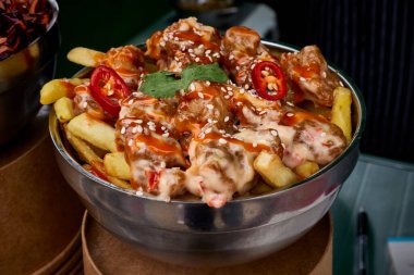 Kore tavuğuyla dolu bir metal kasenin yakından görüntüsü. Patates kızartması, sos ve kırmızı biber serpiştirilmiş. Susam tohumlarıyla süslenmiş.