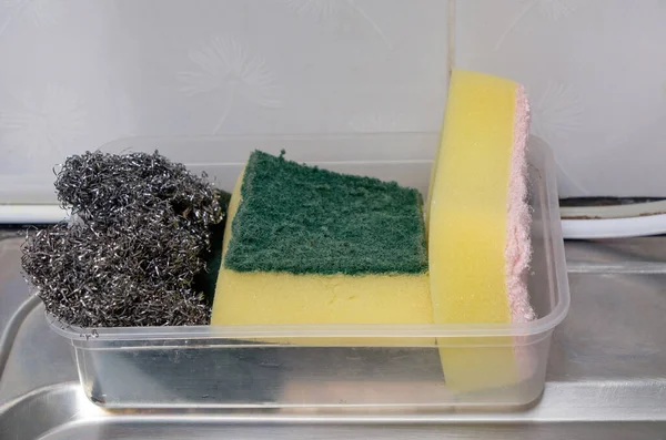 The dishwashing sponge and metal pot scrubber are in the plastic tray for keeping after use, near the edge of the sink in the kitchen, font view for the copy space.