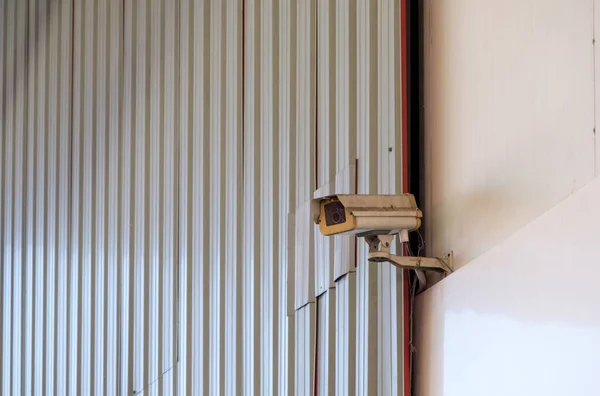 The dirty security video camera is hanging in a high position near the entrance of the warehouse in the industrial park, front view for the background.
