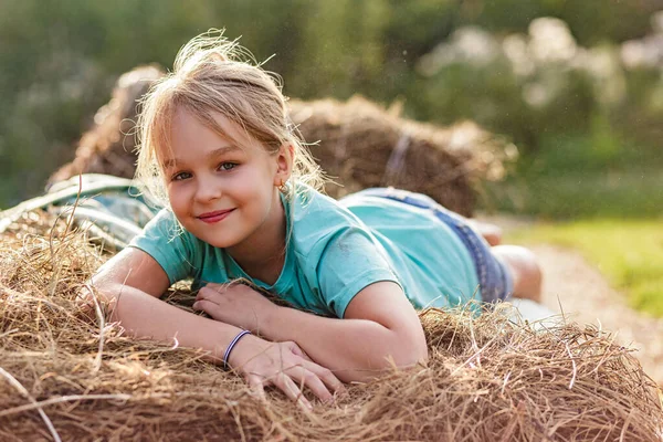 Portrait of a smiling 9-year-old blonde girl lying on a haystack. Happy childhood. A hot summer evening. — Stock Photo, Image