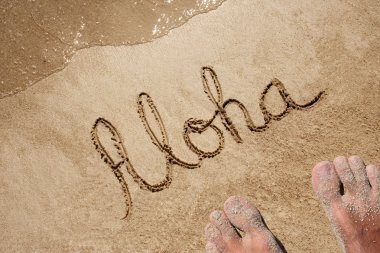 Concept or conceptual Aloha text handwritten in sand on a beach with feet in an exotic island for tropical