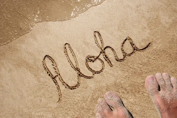 Concept or conceptual Aloha text handwritten in sand on a beach with feet in an exotic island for tropical — Zdjęcie stockowe