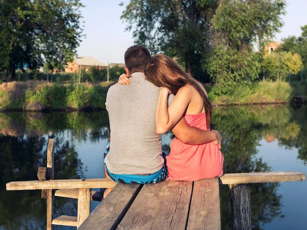 Jovem casal sentado abraçando na ponte junto ao rio — Fotografia de Stock