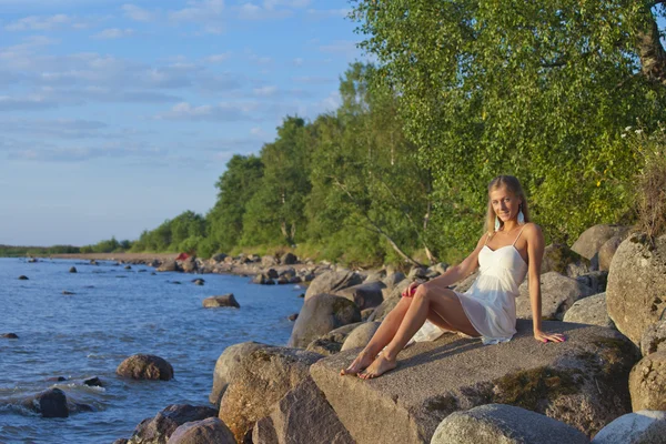 Chica joven en vestido blanco sentado en las rocas en la orilla del lago — Foto de Stock