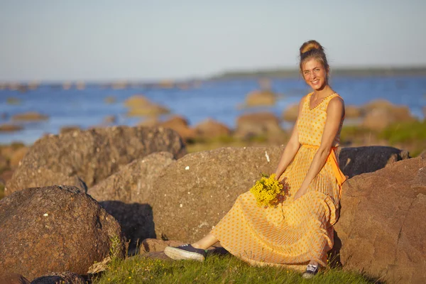 Niña en un vestido amarillo sentado en las rocas con un ramo de flores — Foto de Stock