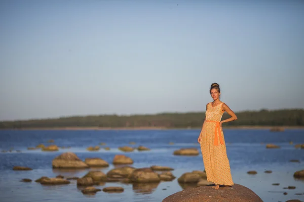 Ung flicka i en gul klänning står på stenar på stranden vid viken — Stockfoto