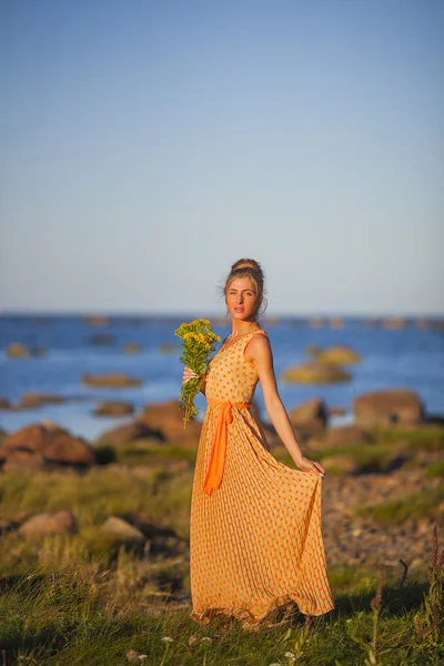 Ung flicka i en gul klänning står på stenar på stranden vid viken — Stockfoto