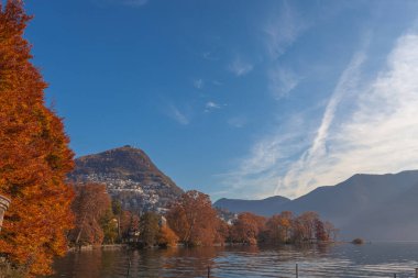 Parco Ciani göl kenarındaki sonbahar sabahı manzarası, turuncu ve kırmızı sonbahar renklerinde ağaçlar. Arka planda Bre Dağı var. Lugano, Ticino, İsviçre