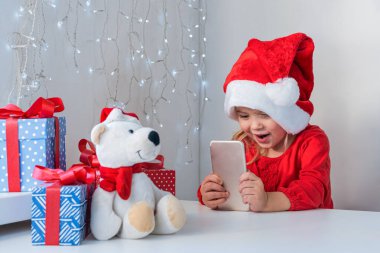Noel Baba şapkalı küçük kız gülümsüyor ve telefonda konuşuyor. Çevrimiçi Noel tebrikleri.