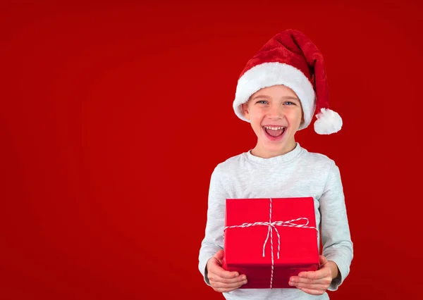 Мальчик в шляпе Санты держит подарок на красном фоне и улыбается. — стоковое фото