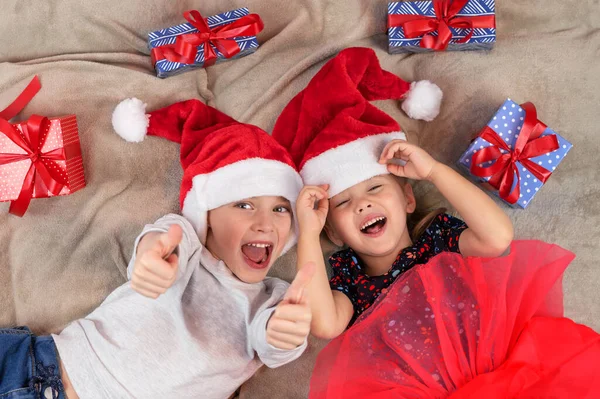 Дети лежат на полу и наслаждаются подарками. Мальчик в рождественской шляпе показывает, как жест, держит большой палец вверх, говорит, что все лучше, рекомендует что-то. — стоковое фото