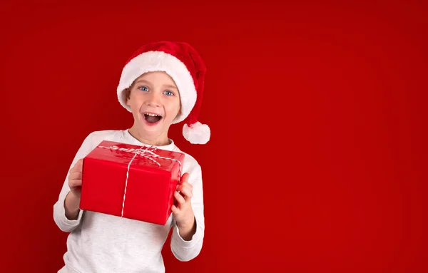 Мальчик в шляпе Санты держит подарок на красном фоне и сюрприз. — стоковое фото