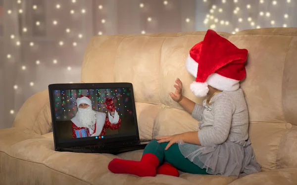 Парень в шляпе Санты сейчас в чате с Сантой. Маленькая девочка слушает Санта-Клауса онлайн. — стоковое фото