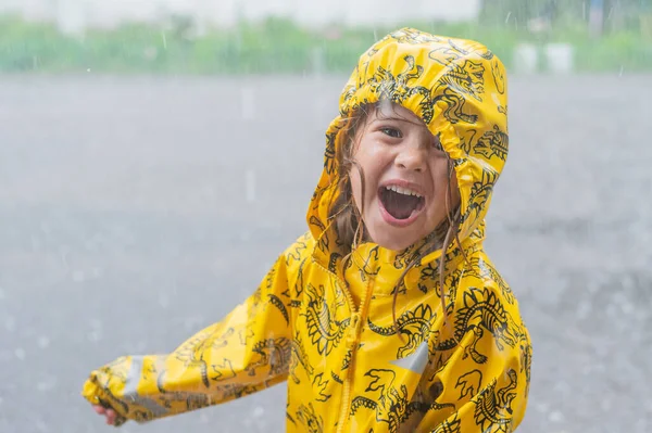 Das Mädchen trägt einen gelben Regenmantel und genießt den Regen. Glückliches Kind unter der Sommerdusche. — Stockfoto