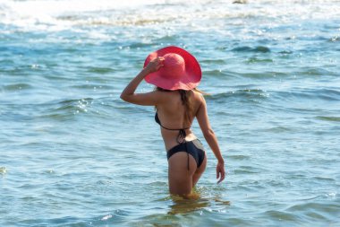 Siyah bikinili ve kırmızı şapkalı, tropik bir plajın sularına giren seksi bir kadının arka görüntüsü.