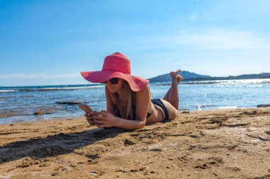 Siyah bikinili ve kırmızı şapkalı bir kadın tropikal plajda uzanmış cep telefonunu güneşlenirken izliyor.