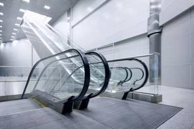 Two sleek, glass and metal escalators rise dramatically toward a brightly lit, modern hall ceiling.