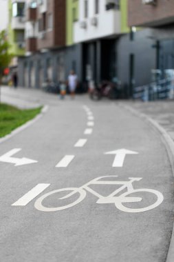 Oklarla ve kesik çizgilerle işaretlenmiş bisiklet şeridine yakın plan, bulanık konut binası ve arka plandaki insanlar.
