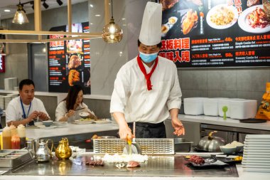 Şanghay, Çin - 16 Mayıs 2025 'te bir restoranda teppanyaki usulü yemek hazırlayan bir şefi yakalıyor, şef beyaz bir üniforma, uzun bir şapka ve yüz maskesi takıyor.